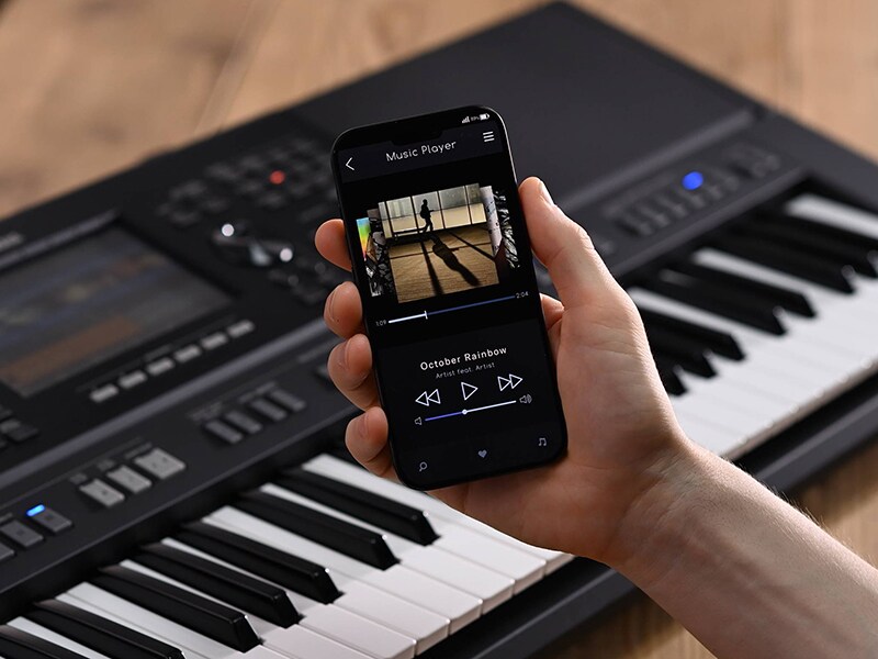 Estación de trabajo de arreglos Yamaha PSR-SX920 y una mano sosteniendo un smartphone con la pantalla de un reproductor de música.
