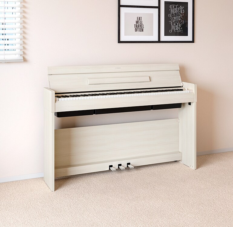 Una sala de estar que cuenta con un piano ARIUS color abedul blanco colocado junto a estantes de pared en los que se encuentran lámparas y un difusor, armoniosamente integrado con la decoración.